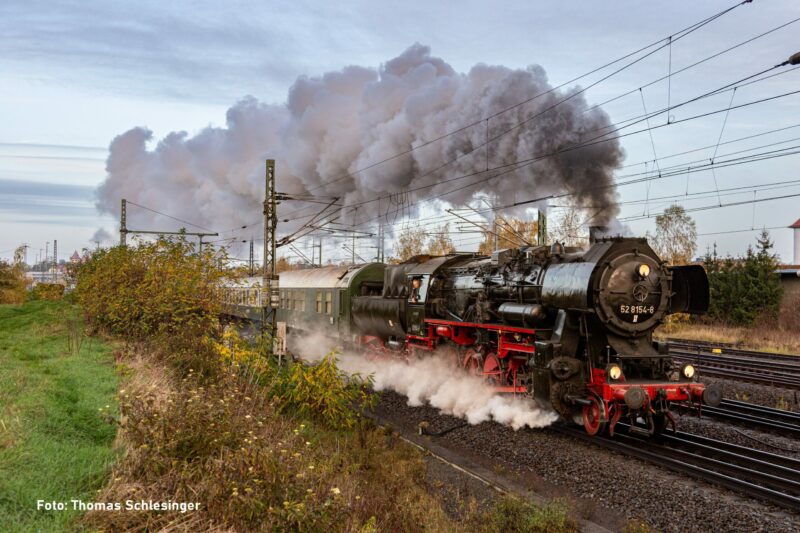 Das Bild zeigt die Dampflok 52 8154 des Eisenbahnmuseums Leipzig mit ausgeprägter Dampffahne, welche sich über den Zug legt. Die Lanschaft ist herbstlich.