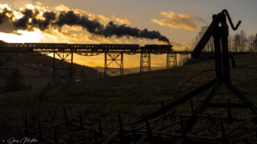 Unser Zug mit langer Dampffahne im Gegenlicht auf dem Markersbacher Viadukt. Im Vordergrund eine Egge eingebettet in die typisch erzgebirgische Hügellandschaft.